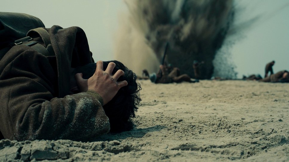 Film still from Dunkirk (2017) showing a young soldier shielding his head as he lies on the ground in front of an exploding bomb