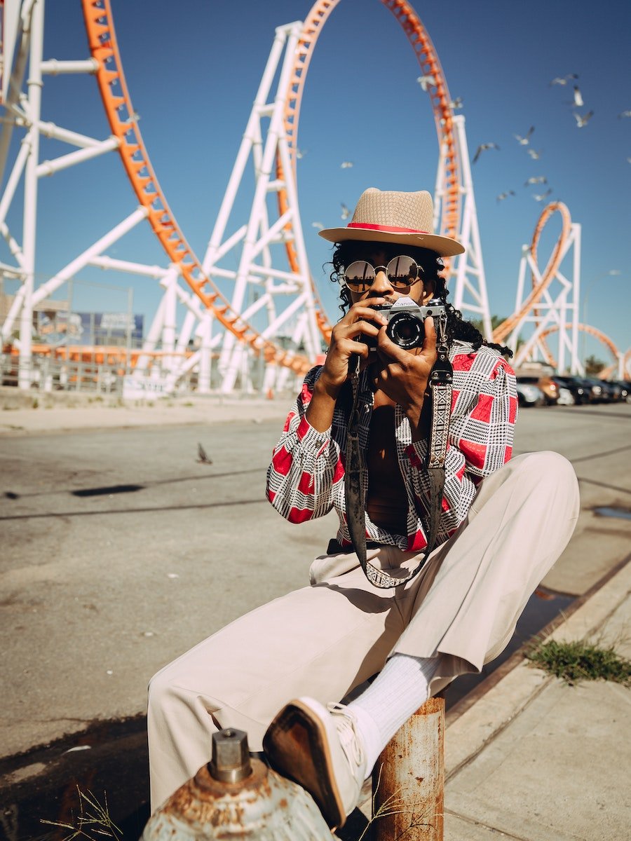 Photographer with film camera seated on a barrier in front of a roller coaster fairground, scouting urban filming locations